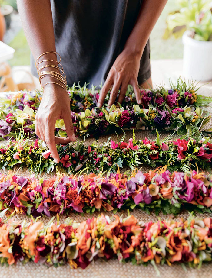 Row of lei on a straw mat with focus on a person's hands wearing gold bangles